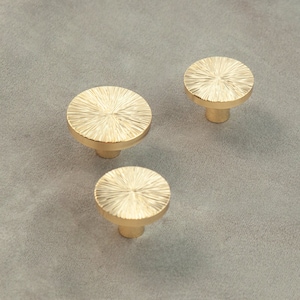 May include: Three gold-toned round cabinet knobs with a textured, sunburst design. The knobs have a short, cylindrical base.