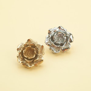 May include: Two decorative floral knobs. One is gold-toned, the other is silver-toned. Each knob is shaped like a blooming rose with detailed petals. The knobs are likely for furniture or decorative purposes.