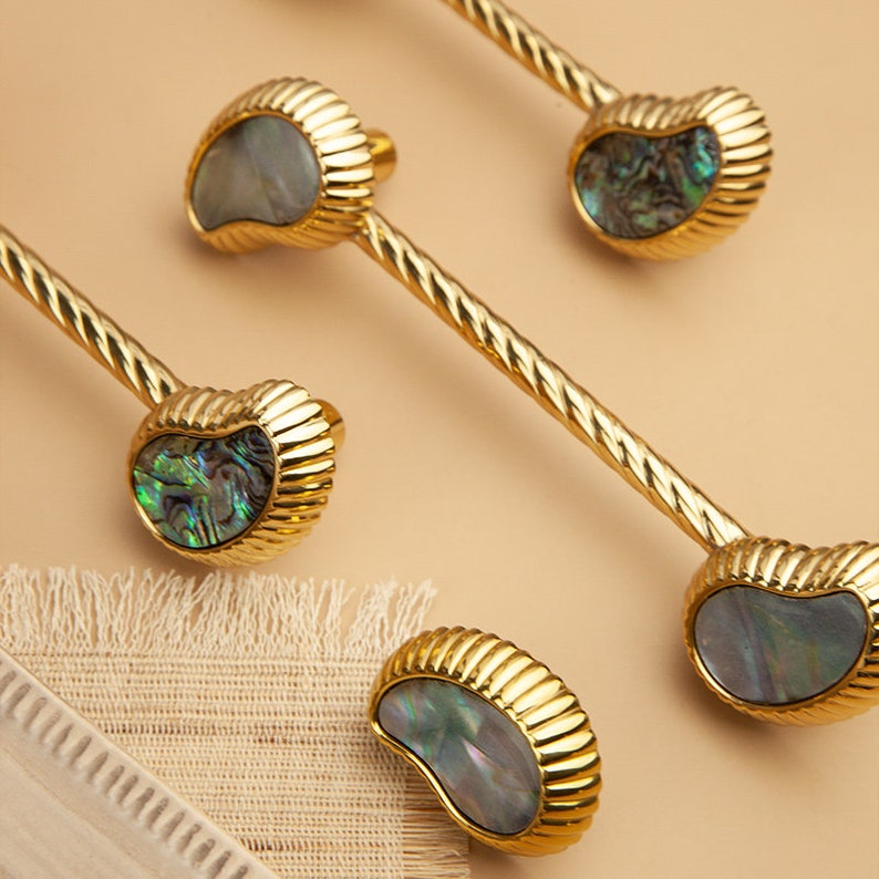 May include: Five gold cabinet knobs with a rope-like design and iridescent abalone shell insets. The knobs are arranged in a staggered pattern on a light brown background.