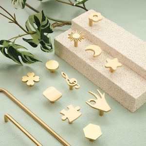 May include: A collection of brass-colored cabinet knobs in various shapes, including a cloud, sun, moon, star, circle, clover, musical note, square, puzzle piece, hand gesture, and hexagon. Two long, brass-colored handles are also present.