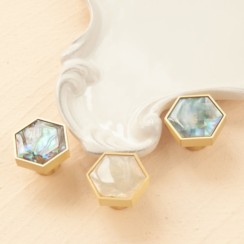 May include: Three hexagonal cabinet knobs with a gold finish and iridescent mother-of-pearl inlays. The knobs are arranged on a white surface.