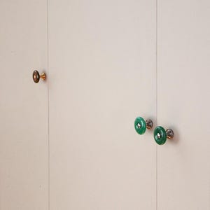 Puede incluir: Tres pomos circulares para armarios fijados sobre una superficie clara. Un pomo es de color marrón oscuro con base dorada, mientras que los otros dos son de color verde vibrante con bases plateadas. Cada pomo tiene un orificio central y acabado liso.