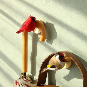 Op de afbeelding: Twee houten vogelhaken aan een muur bevestigd. De haken zijn gevormd als een kardinaal en een goudvink. De kardinaal is rood en de goudvink is geel, bruin en wit.