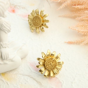 Op de afbeelding: Een paar gouden zonnebloem oorbellen. De oorbellen zijn gemaakt van metaal en hebben een gedetailleerd ontwerp.
