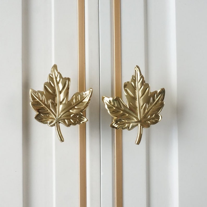 Puede incluir: Dos tiradores de puerta de armario en forma de hoja de arce de color dorado. Las hojas son detalladas y tienen un acabado ligeramente texturizado.