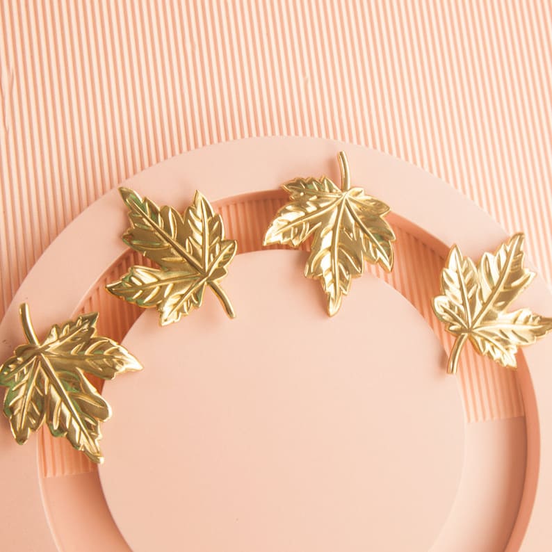 Puede incluir: Cuatro adornos de hoja de arce dorados sobre un fondo de color melocot&oacute;n. Las hojas est&aacute;n dispuestas en c&iacute;rculo alrededor de un espacio en blanco en el centro.