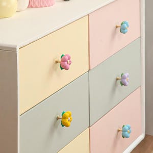 Könnte beinhalten: Eine mehrfarbige Kommode mit sechs Schubladen. Jede Schublade ist in einer anderen Pastellfarbe lackiert und hat einen blütenförmigen Knauf. Die Knöpfe sind in Rosa-, Gelb-, Blau- und Lilatönen gehalten.