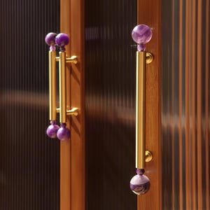 Könnte beinhalten: Schrankgriffe mit goldfarbenen Griffen und lila Edelsteinakzenten. Die Griffe haben ein vertikales Stangendesign mit einer dekorativen Kugel oben und unten. Die Schranktüren sind aus Holz.