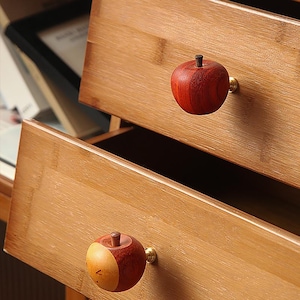 May include: Two wooden drawer pulls shaped like apples. The top pull is red and the bottom pull is red and yellow. Both pulls have a gold metal knob.