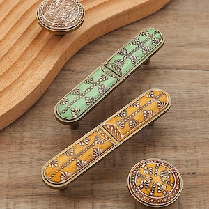 May include: Decorative cabinet hardware featuring two rectangular pulls and two round knobs. The pulls are green and yellow with intricate patterns, while the knobs have a similar design. All pieces have a bronze-colored metal frame and are set against a wooden background.