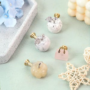 K&ouml;nnte beinhalten: Eine Sammlung dekorativer Flaschenverschl&uuml;sse in verschiedenen Formen und Farben. Die Verschl&uuml;sse umfassen Marmor-Apfel-Designs mit silbernen Blattakzenten, eine hellrosa Handtasche und einen gelben K&uuml;rbis. Die Verschl&uuml;sse haben goldfarbene Metallstiele.