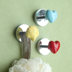 Op de afbeelding: Drie decoratieve haken met een chromen afwerking en keramische accenten. De haken zijn gevormd als een hart, een schelp en een ander hart. De haken zijn gemonteerd op een lichtgroene muur.