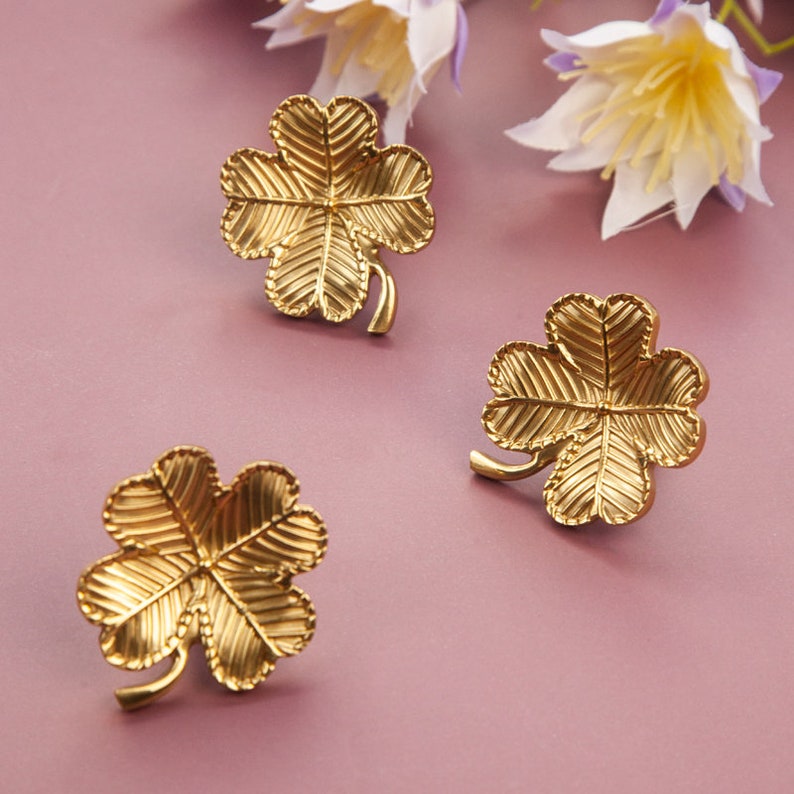 Peut inclure: Trois breloques tr&egrave;fles &agrave; quatre feuilles de couleur dor&eacute;e avec une surface textur&eacute;e. Les breloques sont sur un fond rose.