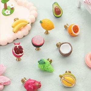 May include: A collection of decorative cabinet knobs in various fruit designs, including bananas, lemons, avocados, and pomegranates. Each knob features a detailed fruit shape with a brass-colored base. The knobs are designed to add a playful and colorful touch to furniture.