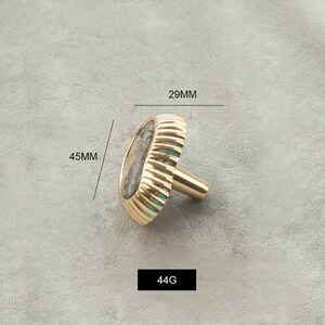 May include: A gold-toned cabinet knob with a decorative, oval-shaped shell inlay. The knob measures 29 mm in diameter and 45 mm in height. It weighs 44 grams.