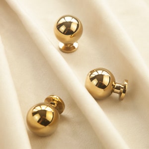 Könnte beinhalten: Drei goldfarbene runde Schrankknöpfe mit glatter, glänzender Oberfläche. Die Knöpfe haben eine runde Basis und eine runde Oberseite.