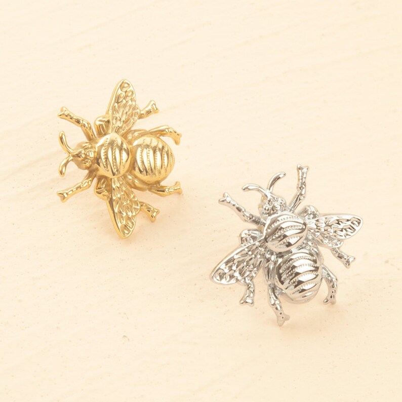 Peut inclure: Deux poign&eacute;es de tiroir d&eacute;coratives en forme d'abeille, l'une dor&eacute;e et l'autre argent&eacute;e, avec des ailes d&eacute;taill&eacute;es et un corps textur&eacute;.