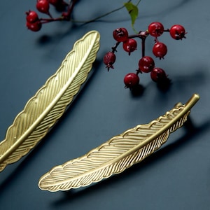 Op de afbeelding: Twee gouden, veer-vormige ladegrepen. De grepen zijn voorzien van een getextureerd veerontwerp.