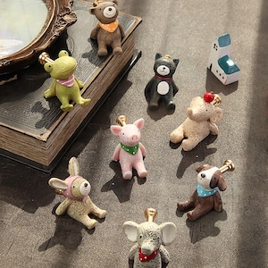 Pode incluir: Uma coleção de pequenas figuras de animais, incluindo um urso, um sapo, um gato, um porco, um elefante e um cão. Cada figura tem um design único com acessórios como bandanas e coroas. As figuras estão dispostas sobre uma superfície escura.