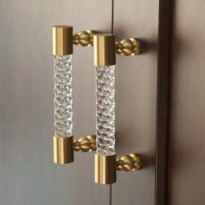 Peut inclure: Une paire de poignées de porte de placard dorées avec des poignées en verre transparent. Les poignées ont un design torsadé.