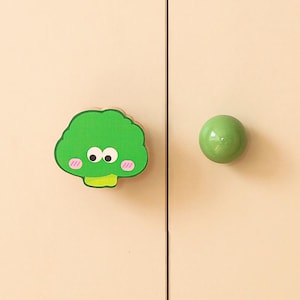 May include: A pair of cabinet knobs. One knob is a round, glossy, light green sphere. The other is a green broccoli-shaped knob with a smiling face, pink cheeks, and a yellow stem. Both knobs are mounted on a light beige cabinet door.