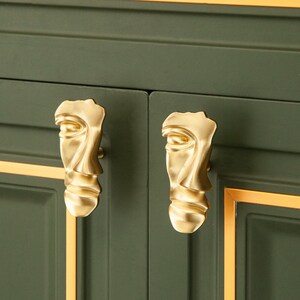 May include: Two gold-toned face-shaped cabinet knobs with a textured surface. The knobs are mounted on a dark green cabinet door with gold trim.