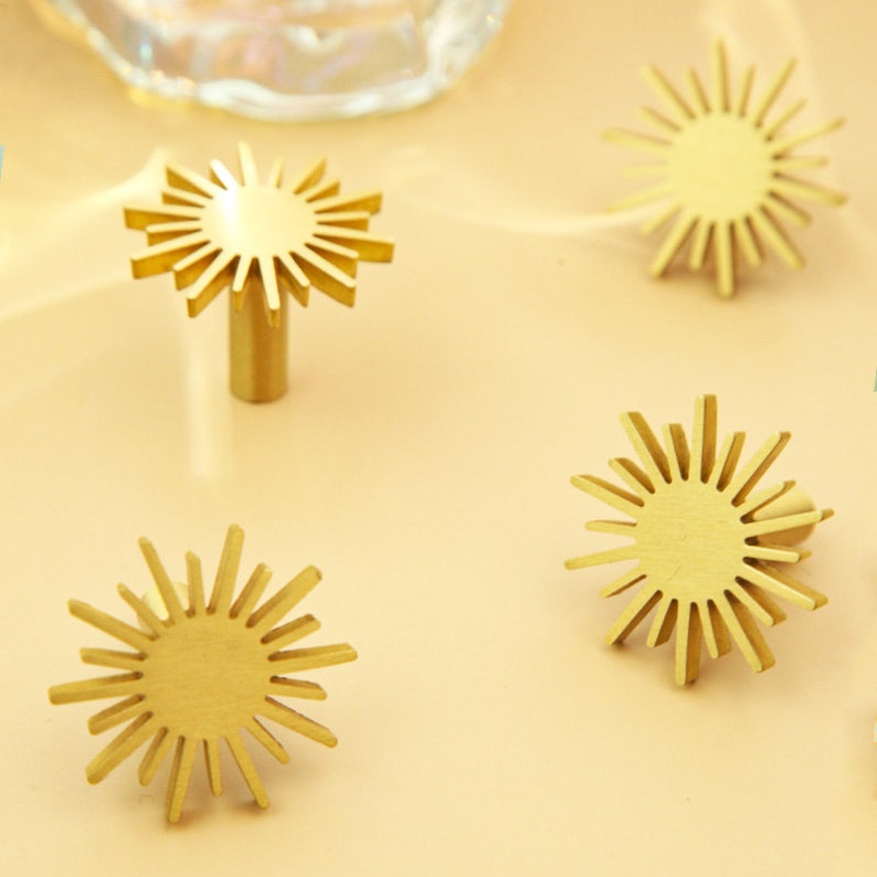 May include: Four gold-toned sunburst-shaped cabinet knobs with a circular center. The knobs are arranged in a square pattern on a light beige surface.