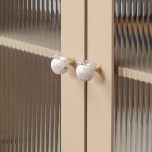 K&ouml;nnte beinhalten: Nahaufnahme einer Schrankt&uuml;r mit zwei apfelf&ouml;rmigen Schrankkn&ouml;pfen. Die Kn&ouml;pfe sind aus wei&szlig;em Marmor mit silbernen Blattakzenten und goldfarbener Hardware. Die Schrankt&uuml;r ist hellbeige.