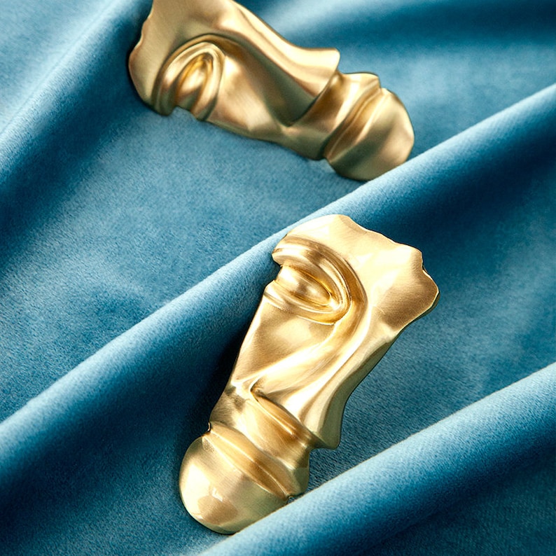 May include: Two gold-colored abstract face sculptures on a blue velvet background.