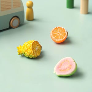 May include: Miniature fruit ornaments, including a pineapple, orange slice, and guava, are displayed on a light green surface. The pineapple is yellow and green, the orange is bright orange, and the guava is pink and green.
