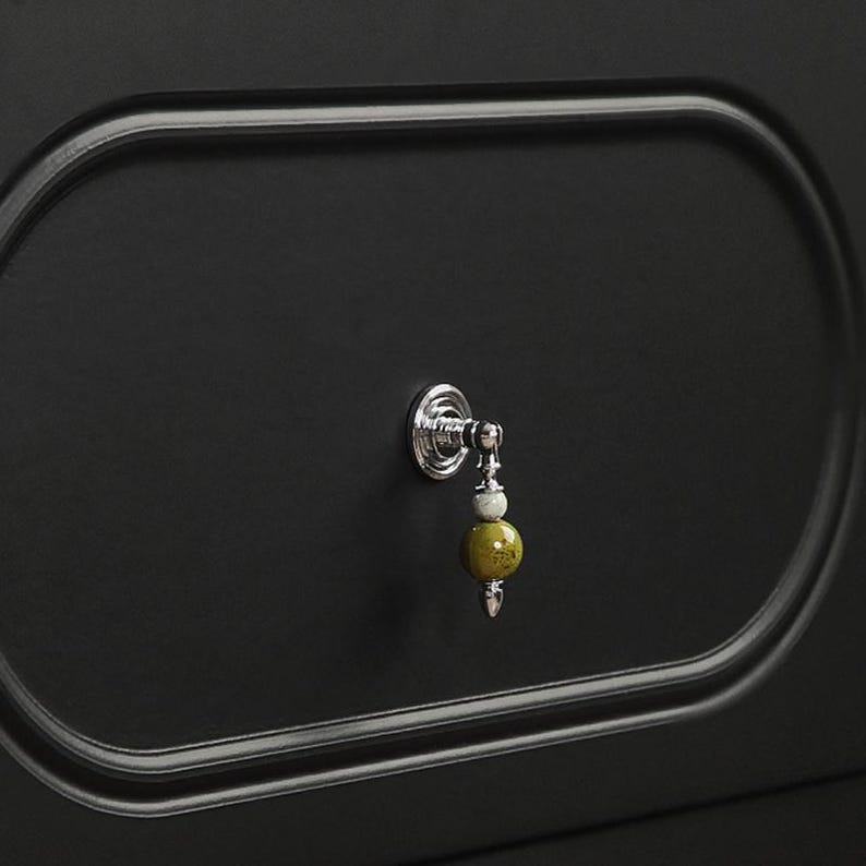 Puede incluir: Primer plano de una puerta de armario negra con un marco ovalado plateado y un tirador decorativo. El tirador presenta una base plateada con un dise&ntilde;o colgante de cuentas amarillas y blancas.
