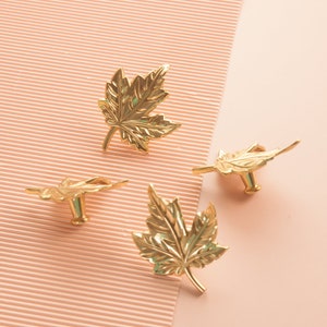 Puede incluir: Cuatro pomos de armario en forma de hoja de arce de color dorado. Los pomos est&aacute;n sobre un fondo de rayas rosas y blancas.