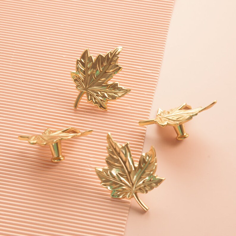 Puede incluir: Cuatro pomos de armario en forma de hoja de arce de color dorado. Los pomos est&aacute;n sobre un fondo de rayas rosas y blancas.