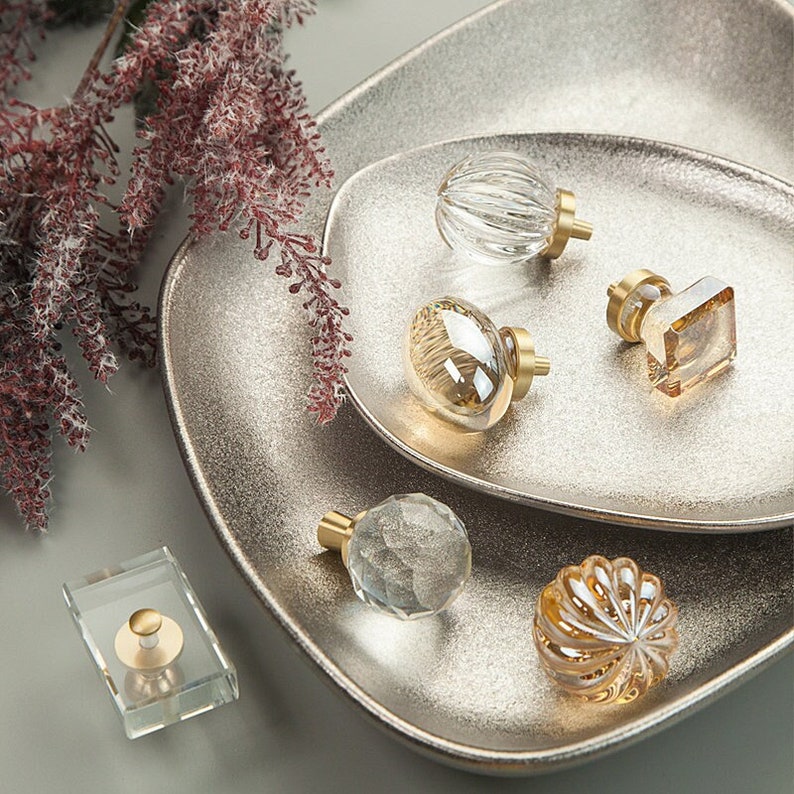 May include: Assortment of six glass cabinet knobs with gold metal bases. The knobs are displayed on a silver tray. The knobs have various shapes and textures, including round, oval, and diamond.