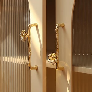 May include: Two gold-colored cabinet handles. Each handle has a branch-like design with a flower at the top. The handles are mounted on a light-colored cabinet door. The background has a textured glass panel.