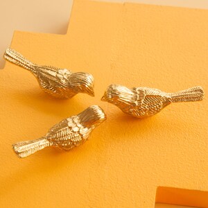 May include: Three gold bird figurines on a yellow background. The birds are facing each other and have detailed feather patterns.