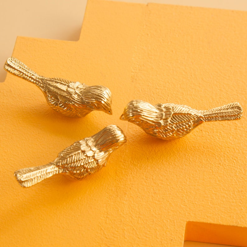 May include: Three gold bird figurines on a yellow background. The birds are facing each other and have detailed feather patterns.