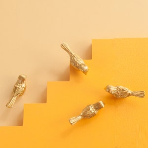 May include: Four gold bird-shaped cabinet knobs on a yellow background. The knobs are detailed with textured feathers and are arranged in a staggered pattern.