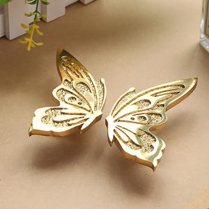 May include: Two gold-coloured butterfly-shaped decorative hardware pieces. Each wing features an intricate, textured pattern. The hardware is likely for furniture or decorative purposes, adding a touch of elegance.