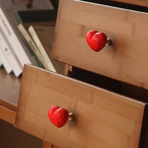 Op de afbeelding: Close-up van twee houten lades met rode hartvormige knoppen. De knoppen zijn getextureerd en bevestigd met goudkleurige hardware. De laden zijn gemaakt van lichtbruin hout.