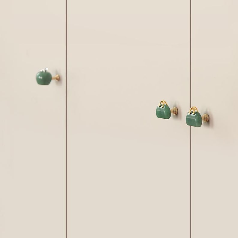 K&ouml;nnte beinhalten: Schrankkn&ouml;pfe in Form kleiner gr&uuml;ner Handtaschen mit goldfarbenen Beschl&auml;gen. Sie sind an einer cremefarbenen Schrankt&uuml;r befestigt. Die Kn&ouml;pfe sind im Abstand angeordnet. Die T&uuml;r ist neutral.