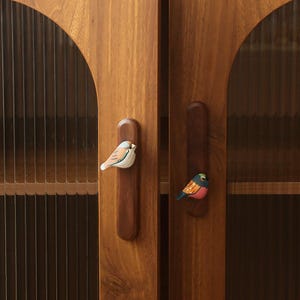 以下が含まれることがあります： 鳥の形をした取っ手が付いた木製のキャビネットドア。キャビネットのドアは温かみのある茶色で、取っ手はカラフルな鳥のデザインで塗装されています。キャビネットには、縦線のあるガラスパネルが付いています。