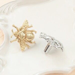 Puede incluir: Dos pomos de armario con forma de abeja, uno dorado y otro plateado, sobre una superficie blanca.