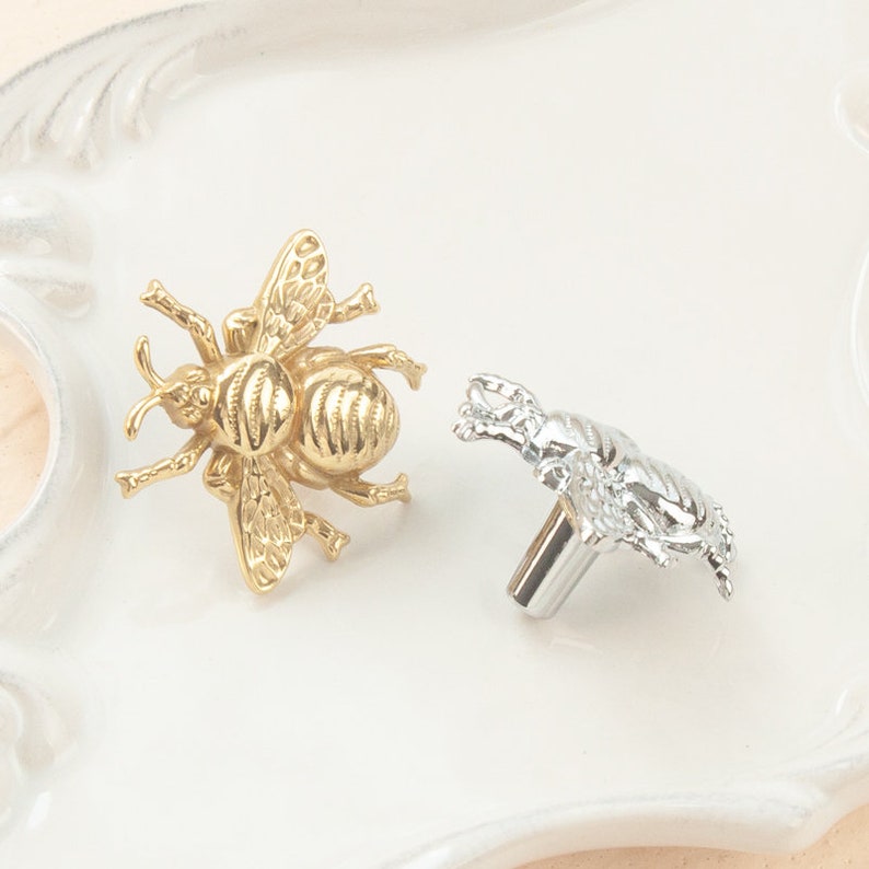 Peut inclure: Deux boutons de porte en forme d'abeille, l'un dor&eacute; et l'autre argent&eacute;, sur une surface blanche.