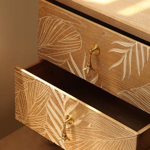 Peut inclure: Tiroirs de commode en bois clair avec un motif de feuilles blanches. Les tiroirs sont orn&eacute;s de boutons en forme de Tour Eiffel dor&eacute;s. Un tiroir est ouvert, r&eacute;v&eacute;lant un int&eacute;rieur sombre.