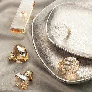 May include: Four decorative glass cabinet knobs with brass bases. The knobs are in various shapes and sizes, including round, oval, and square. The knobs are on a light brown surface.