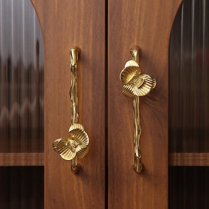 Op de afbeelding: Close-up van twee houten kastdeuren met sierlijke goudkleurige handgrepen. De handgrepen zijn gevormd als bloemen en stelen, wat een decoratief tintje geeft. De kastdeuren zijn warmbruin.