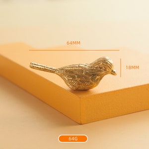 May include: A gold-colored metal bird figurine with detailed wings and a beak. The figurine measures 6.4 cm in length and 1.8 cm in height. It weighs 64 grams.