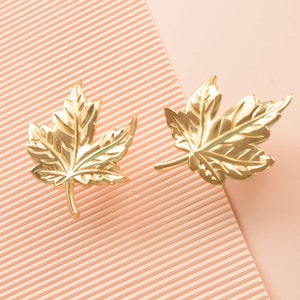 Puede incluir: Un par de pendientes de hoja de arce de color dorado. Las hojas son detalladas y tienen un acabado texturizado.