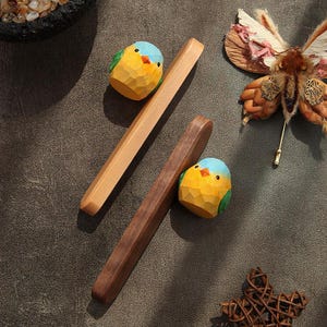 Peut inclure: Deux repose-baguettes en bois avec des figurines d'oiseaux sculptés. Un repose-baguettes est de couleur claire, l'autre est foncé. Les oiseaux sont jaunes, bleus et verts. Les articles sont sur une surface sombre et texturée.